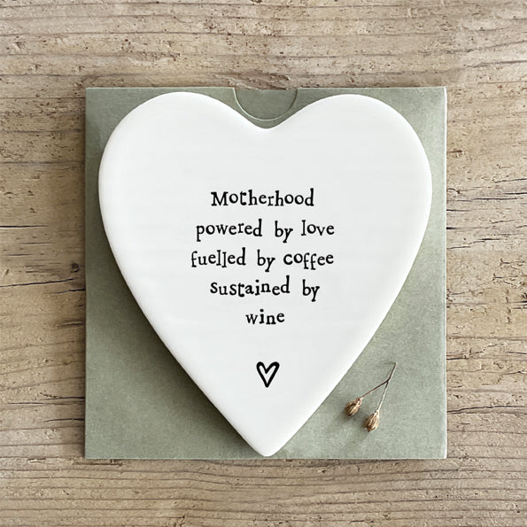 White porcelain heart shaped coaster with black text which reads Motherhood, powered by love, fuelled by coffee, sustained by wine