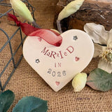 a ceramic heart embossed with lettering that reads married in 2026, a red ribbon is used for hanging the heart up