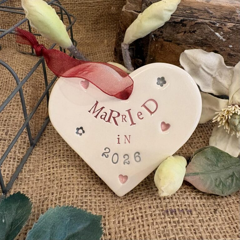 a ceramic heart embossed with lettering that reads married in 2026, a red ribbon is used for hanging the heart up