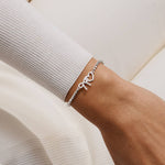 A close-up of a person's wrist wearing a silver beaded bracelet with a bow and heart charm, styled with a ribbed off-white long-sleeve top.
