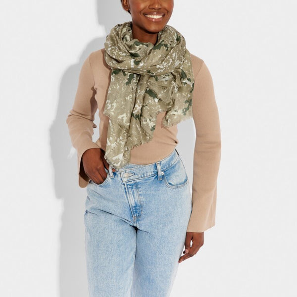 a smiling model in casual clothing wears a scarf in an olive green coloured abstract animal print design with silver details